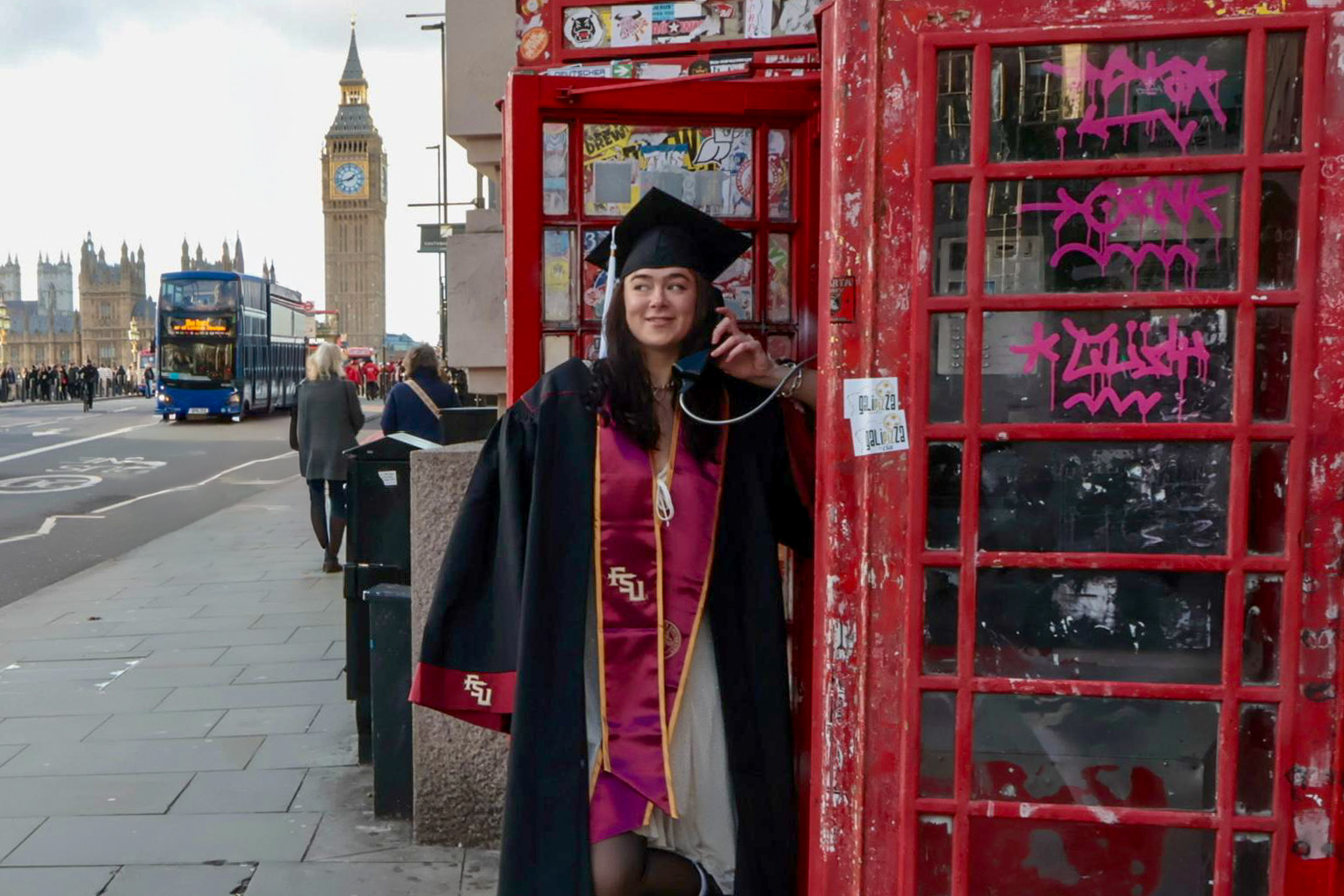 FSU student posing in London in graduation regalia with a London phone booth, holding a phone.