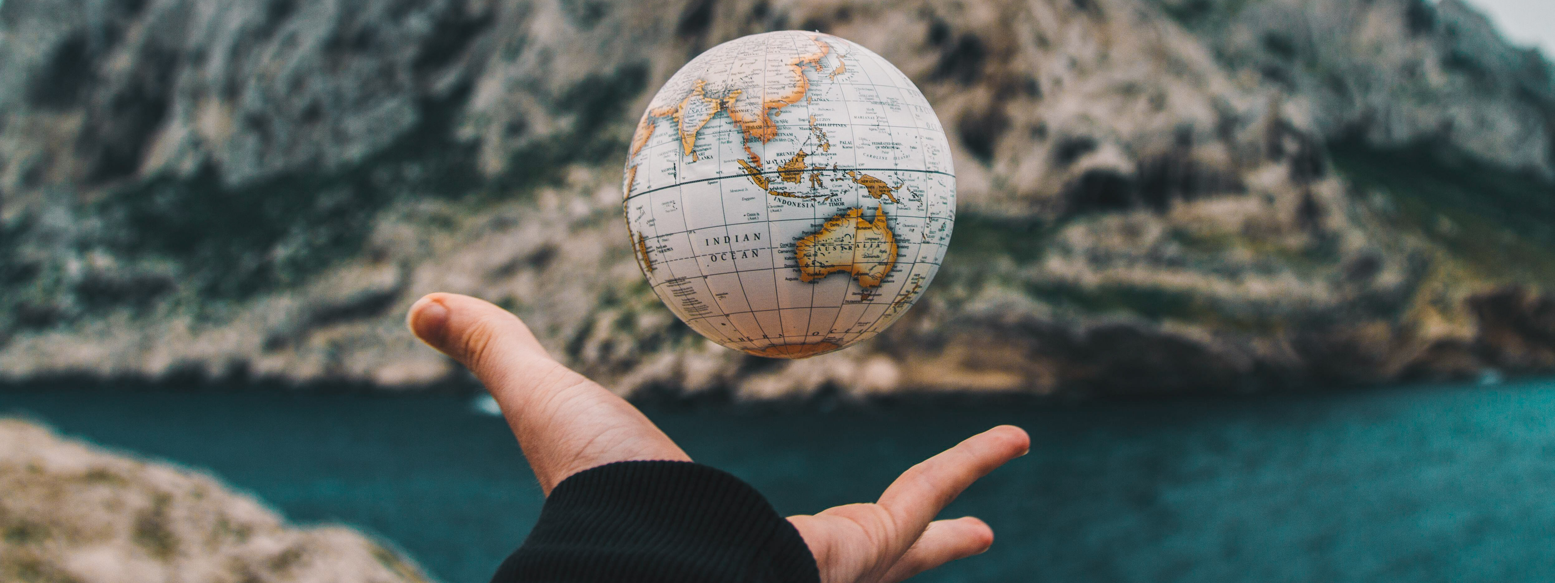 A hand grasping for a small globe with water and rocky mountains in the background.