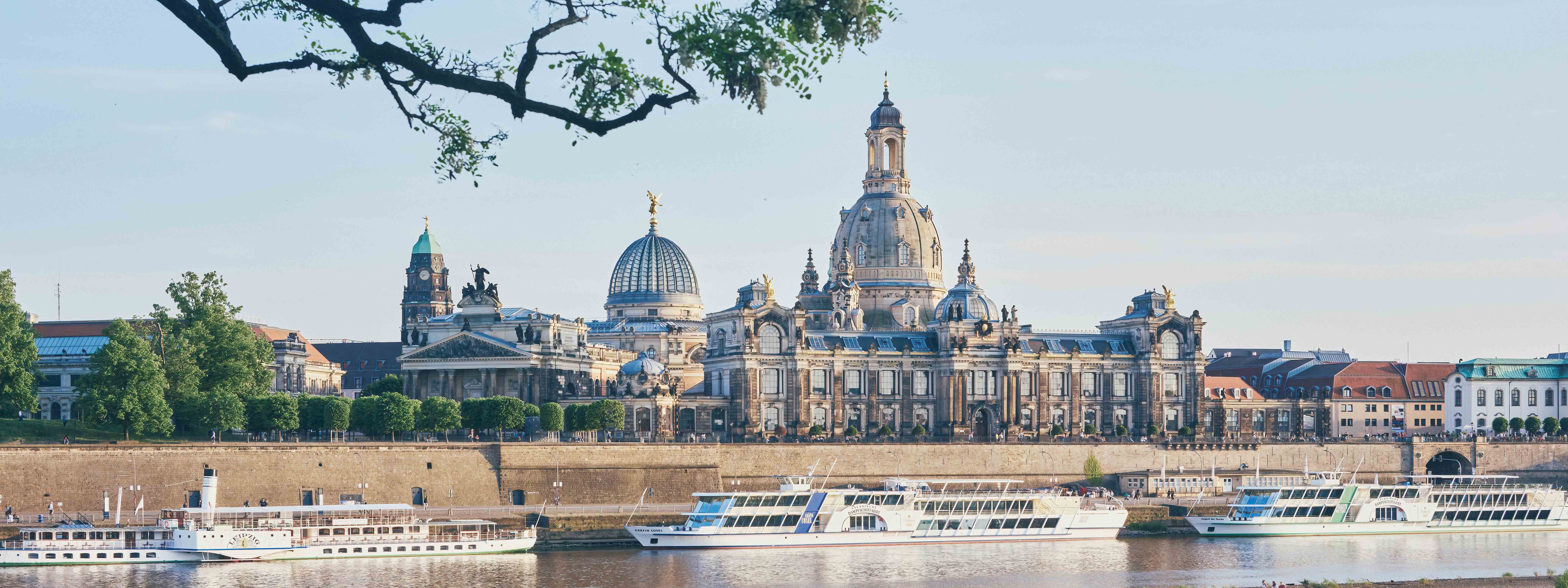 A view of historic buildings in Dresden with a river, boats, and trees in the foreground.