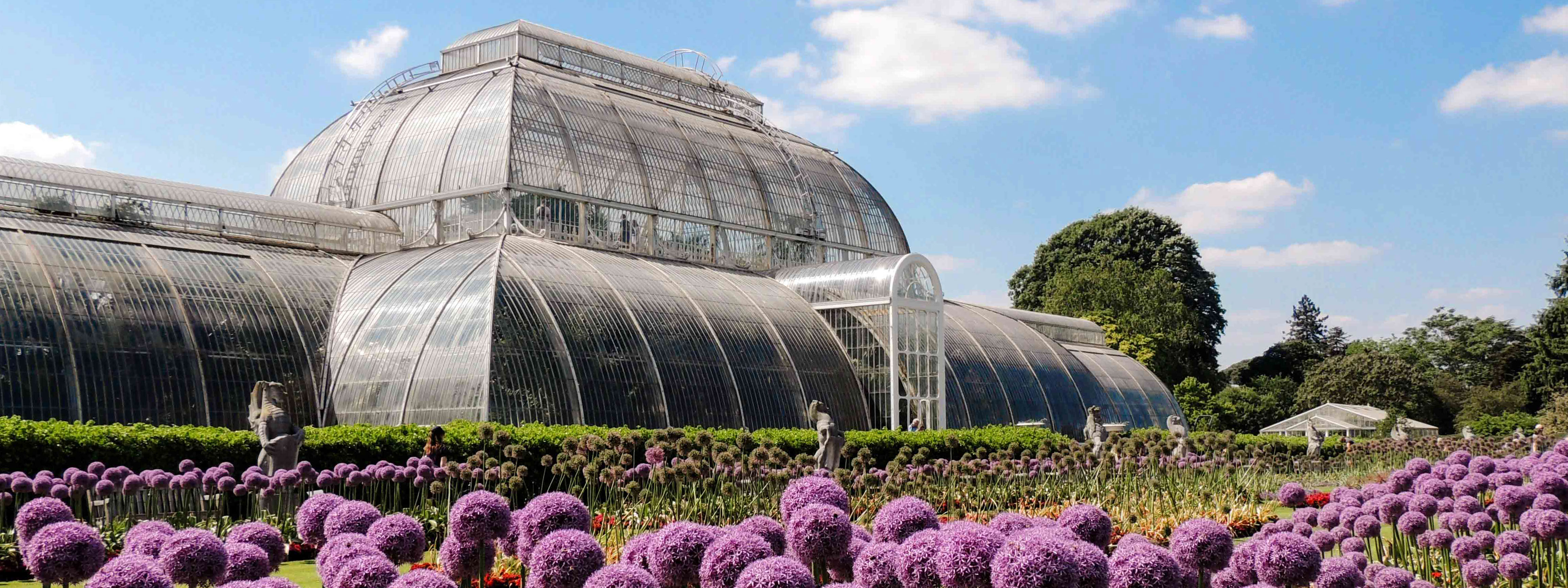 A view of the Kew Gardens greenhouse with flowers and gargoyle statues in the foreground.